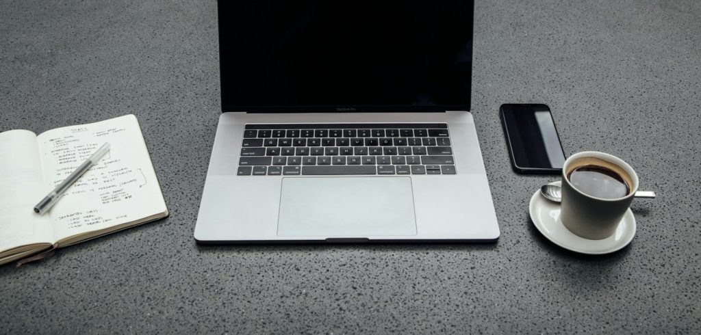 Turned-off MacBook Pro between cup of coffee, iPhone, notebook, and pen