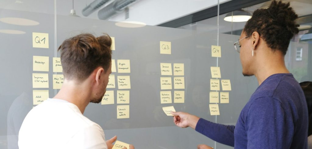 Two people collaborating on a project plan using yellow sticky notes on a glass wall