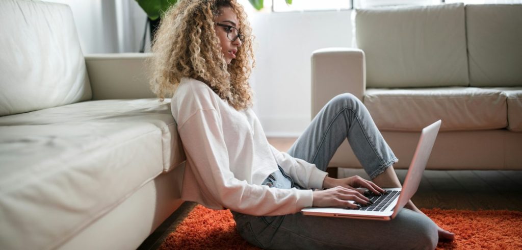 Woman sitting on floor and leaning on couch using laptop